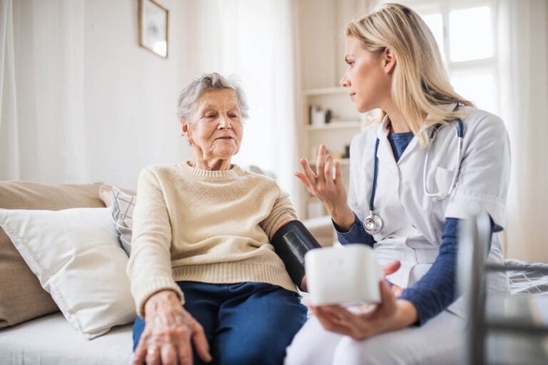 Pflegekraft misst bei einer Seniorin zu Hause den Blutdruck und erklärt ihr die Werte