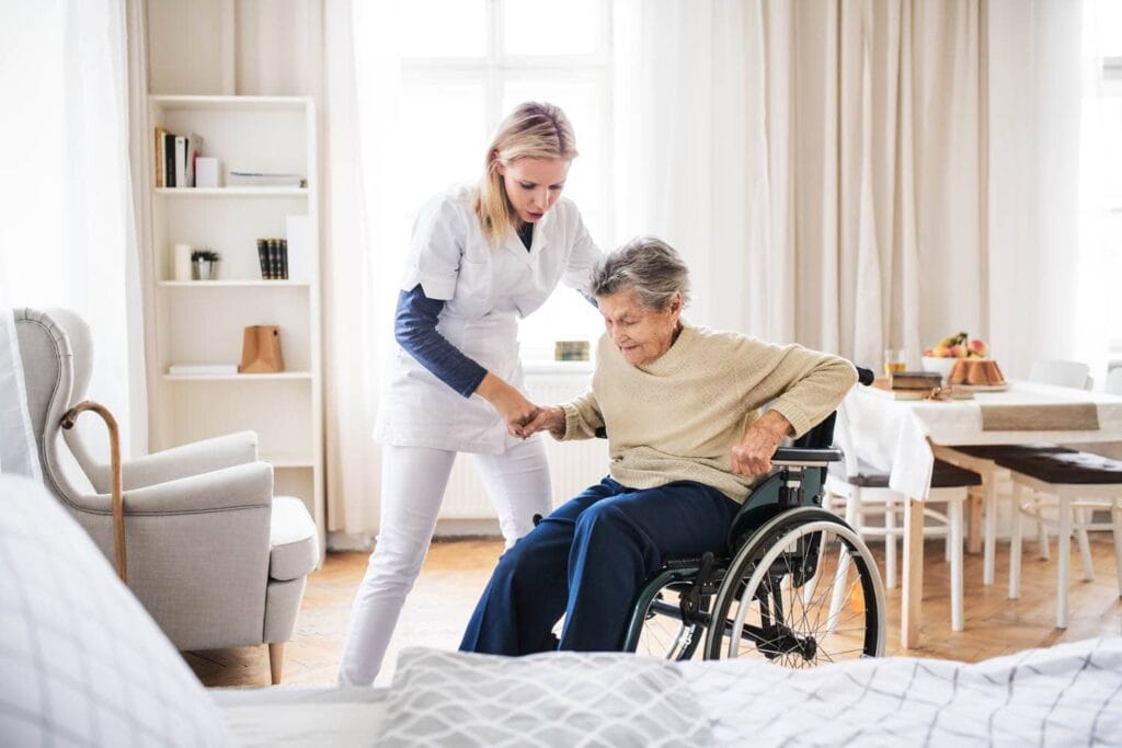 Pflegekraft hilft einer Seniorin beim Aufstehen aus dem Rollstuhl in ihrer Wohnung